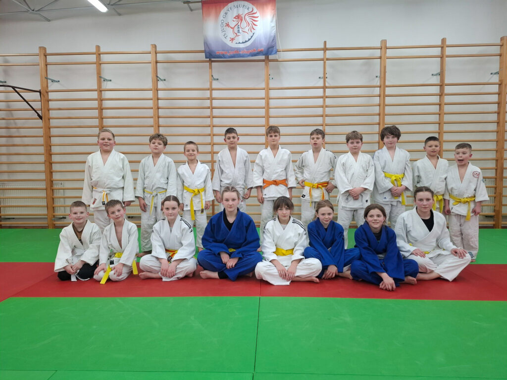 Grupa dzieci z TS Judo Gryf Słupsk w strojach do judo, z uśmiechami pokazująca zdobyte medale i dyplomy podczas turnieju w Koszalinie.