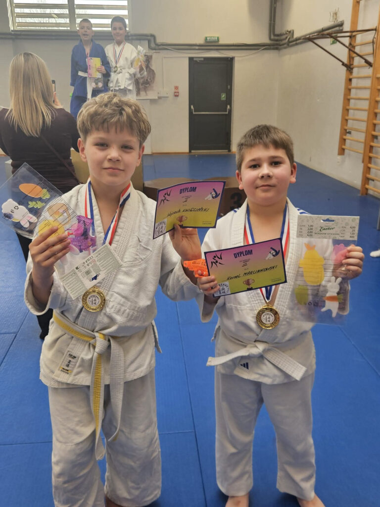 Chłopiec ze Słupska w stroju judo pokazuje swój dyplom i medal na tle drabinek lub trybun w koszalińskiej hali sportowej.