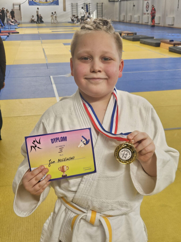 Młody zawodnik TS Judo Gryf Słupsk w białej judodze, uśmiechnięty, trzymający zdobyty medal i dyplom z zawodów w Koszalinie.