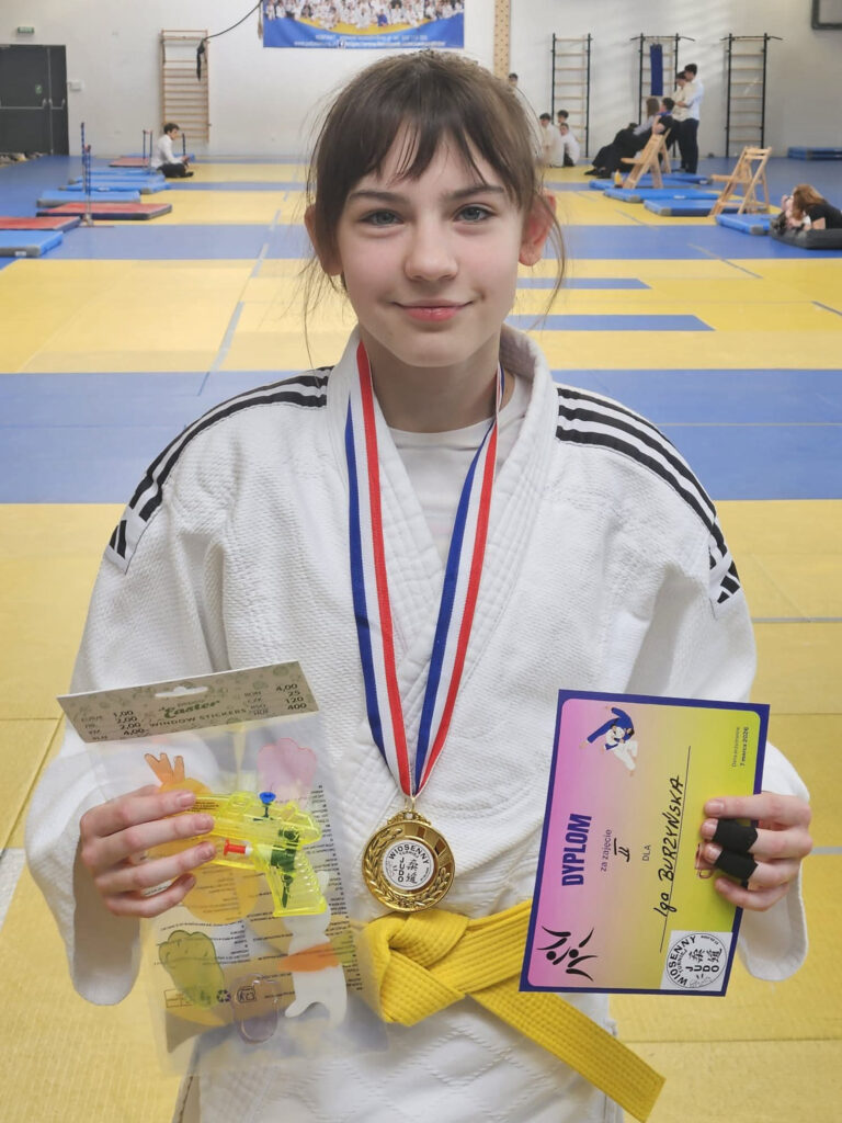 Uśmiechnięta dziewczynka w judodze pokazuje do obiektywu zdobyty dyplom oraz medal z turnieju judo.
