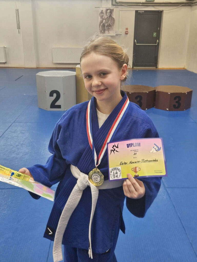 Zbliżenie na dłonie zawodnika w niebieskim kimonie, trzymające pamiątkowy dyplom i błyszczący medal z zawodów w Koszalinie.