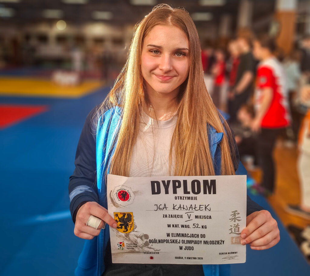 Młoda judoczka w niebieskiej kurtce uśmiecha się, trzymając przed sobą dyplom za zajęcie V miejsca w eliminacjach do Ogólnopolskiej Olimpiady Młodzieży w wadze 52 kg. Tło to rozmyta hala sportowa z tatami.