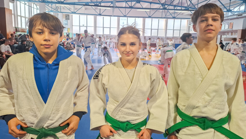 Trójka młodych adeptów judo, jedna dziewczyna z niebieskim dresem pod judogą i dwóch chłopców, stoi w szeregu na niebieskim tatami podczas treningu lub zawodów w hali sportowej.