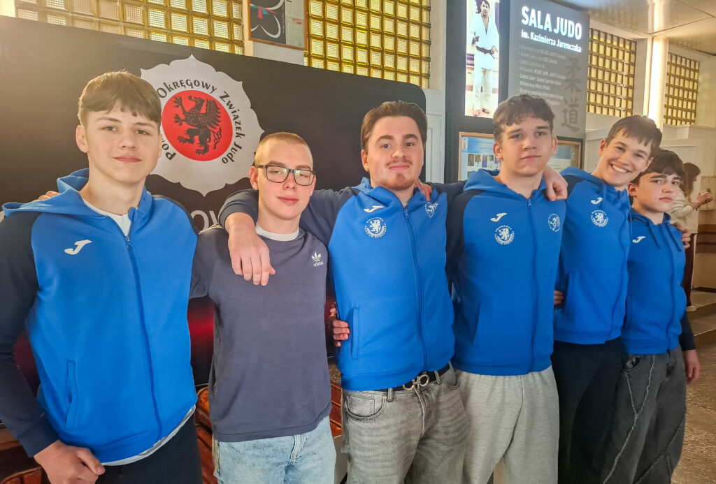 Grupa sześciu młodych sportowców w niebieskich bluzach dresowych z logo TS Judo Gryf Słupsk stoi razem przy ścianie z logotypami. Jeden nosi okulary.