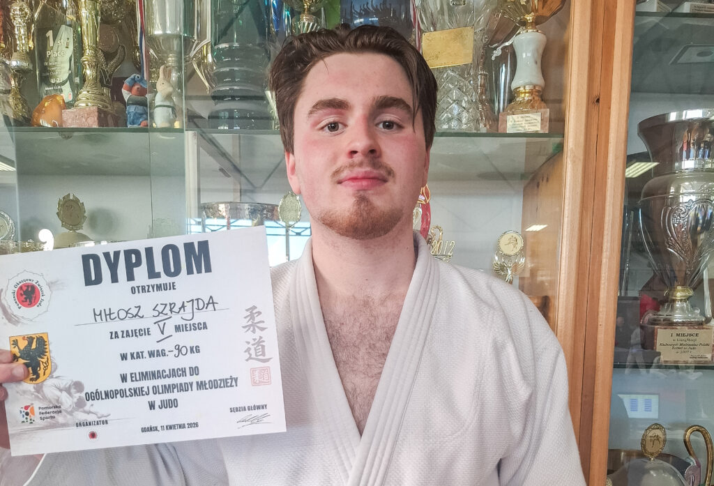 Młody judoka w białych judogach trzyma przed sobą dyplom za zajęcie V miejsca w eliminacjach OOM w wadze 90 kg. Stoi obok szafy wypełnionej sportowymi trofeami.