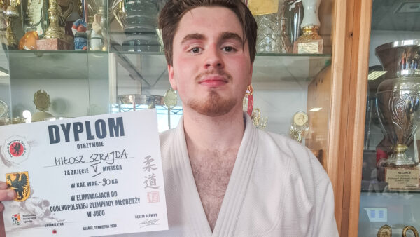 Młody judoka w białych judogach trzyma przed sobą dyplom za zajęcie V miejsca w eliminacjach OOM w wadze 90 kg. Stoi obok szafy wypełnionej sportowymi trofeami.