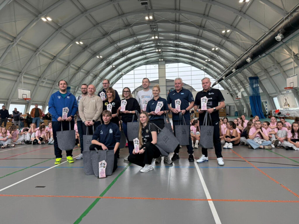 Grupa trenerów i organizatorów Ogólnopolskiego Turnieju Młodziczek w Mielnie pozująca ze statuetkami i pamiątkowymi torbami na tle siedzących na parkiecie zawodniczek.