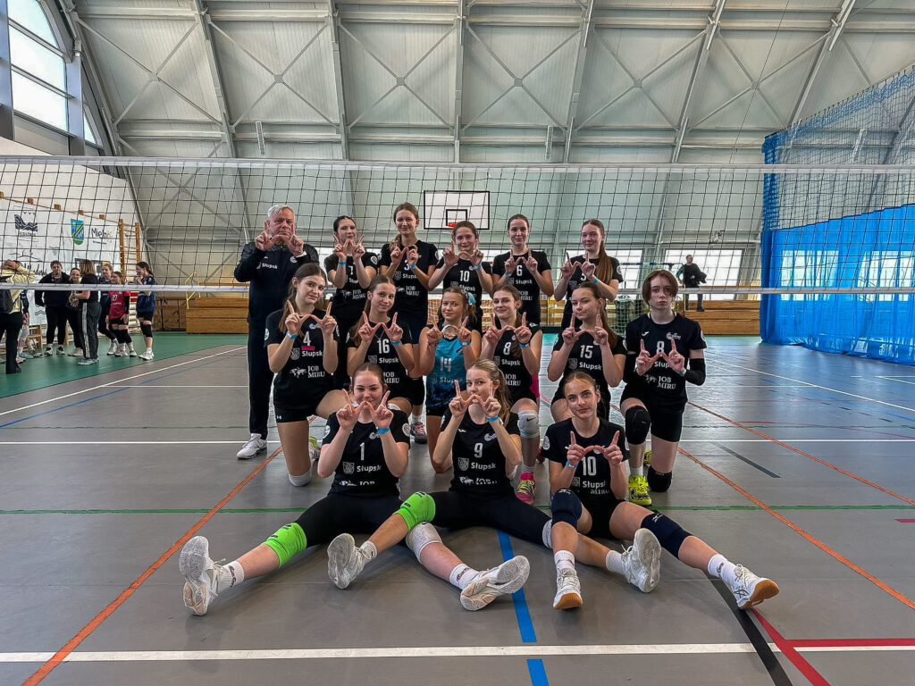 Drużyna młodziczek TPS Czarni Słupsk w czarnych strojach z trenerem, pozująca na jasnym parkiecie w Mielnie i pokazująca uśmiechem radosne gesty dłońmi.