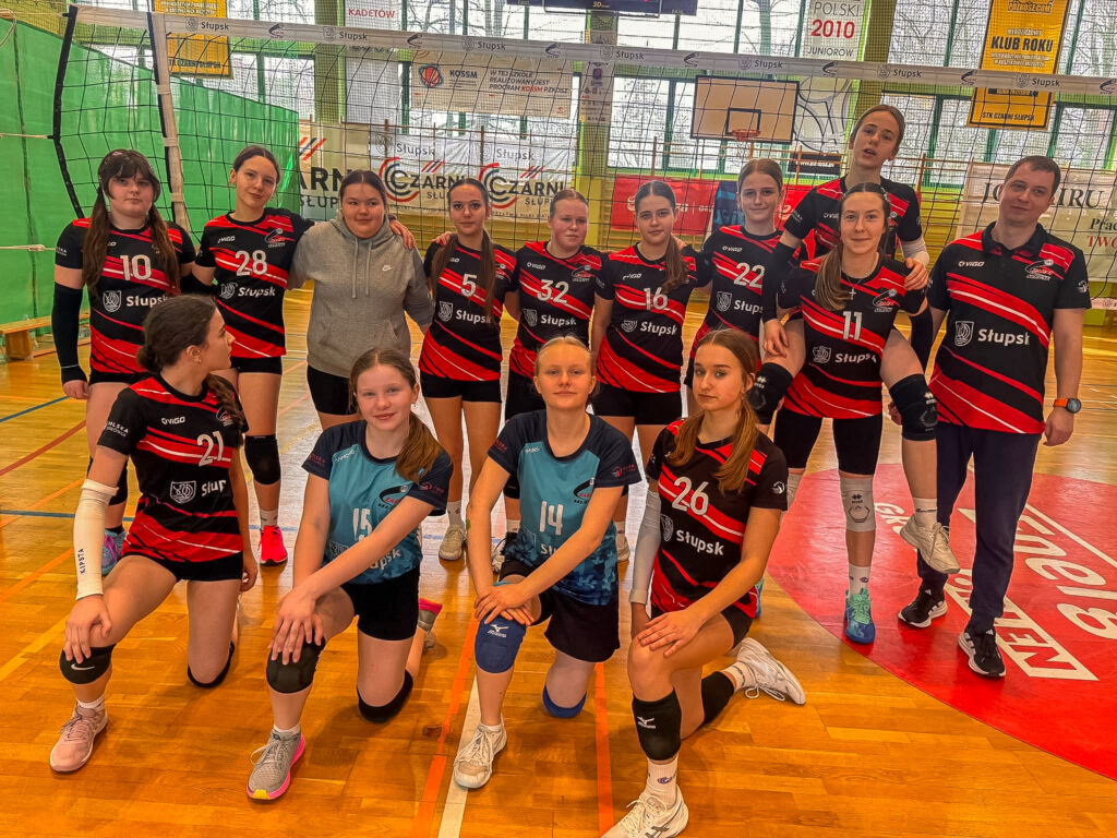 Pamiątkowe zdjęcie drużyny siatkarek TPS Czarni Słupsk wraz z trenerem na hali sportowej. Zawodniczki ubrane są w czarno-czerwone oraz niebieskie stroje meczowe.