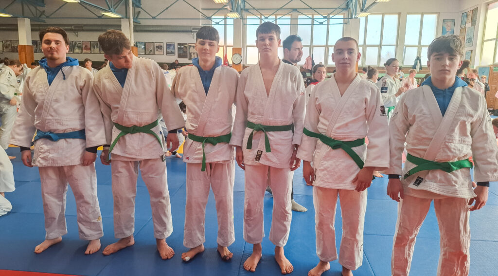 Sześciu młodych judoków w judogach z pasami stoi w szeregu na niebiesko-czerwonym tatami w jasnej hali sportowej. Inni ćwiczący są widoczni w tle.
