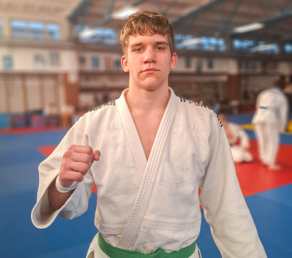 Młody judoka w białych judogach z zielonym pasem zaciska prawą pięść przed sobą i patrzy bezpośrednio w kamerę na tle rozmytej hali sportowej z matami.